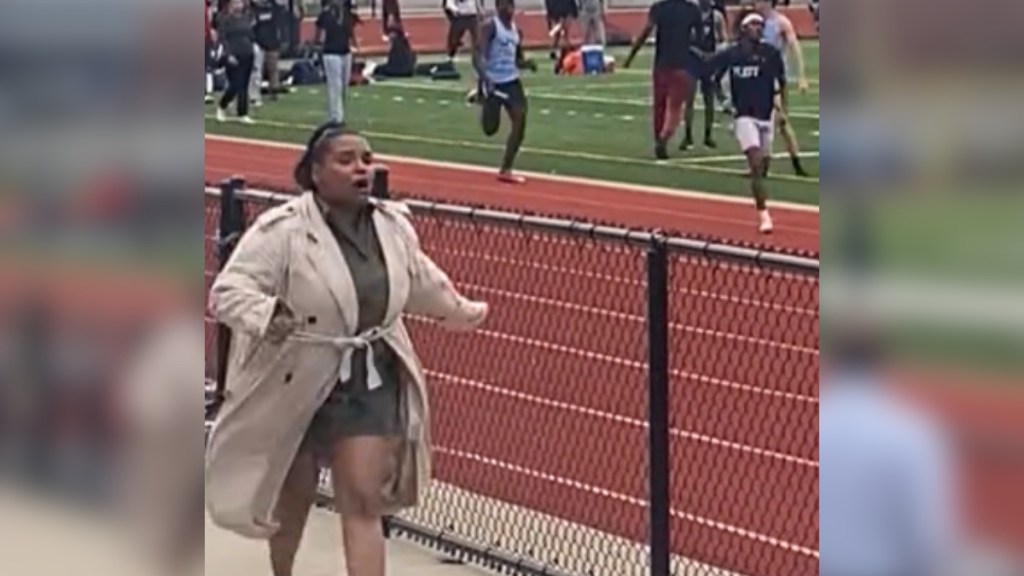 A woman runs on the sidelines of a track field, mouth open because she's speaking.
