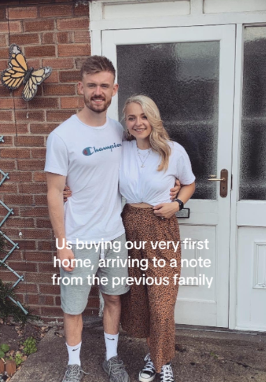 A man and woman smile as the pose outside of their new home at the front door. Text on the image reads: Us buying our very first home, arriving to a note from the previous family