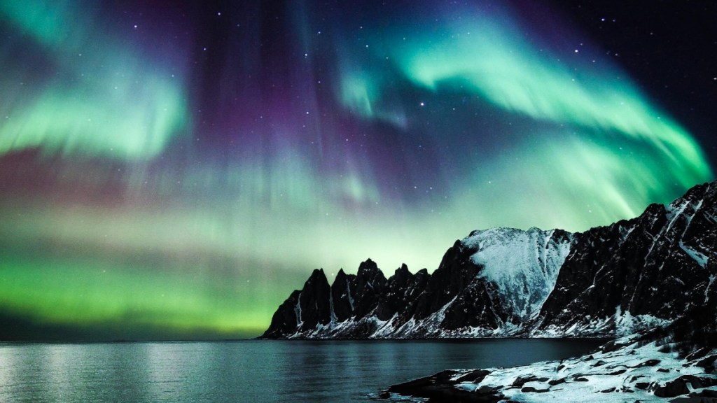 View of the northern lights above snowy mountains and water.