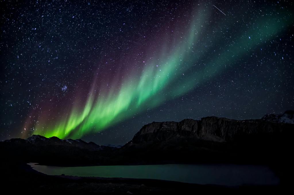 View of the northern lights in a particularly dark area. Shadows of mountains can be seen below.