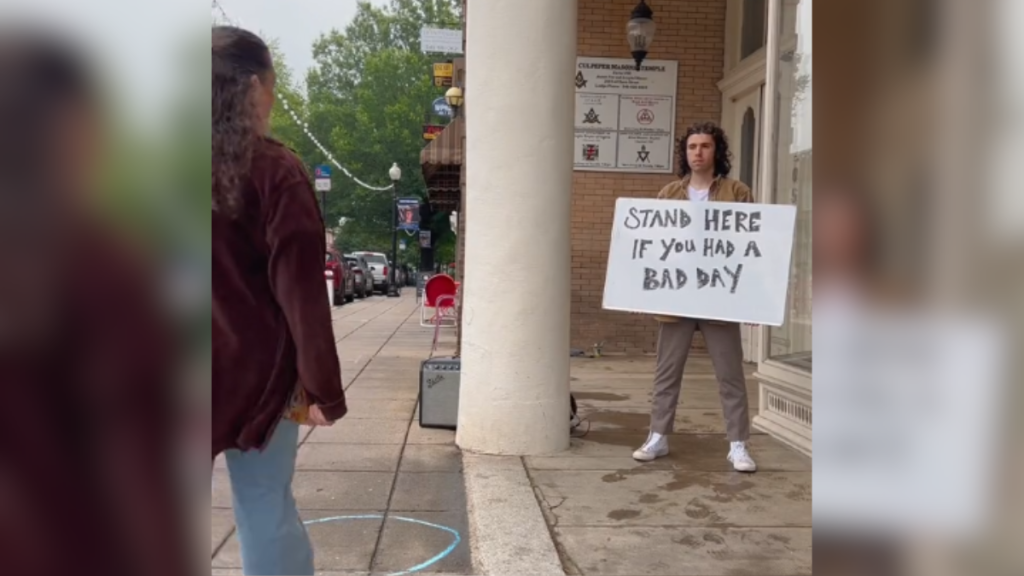 A man holds up a sign that reads "stand here if you had a bad day" as a woman starts to approach him.