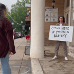 A man holds up a sign that reads "stand here if you had a bad day" as a woman starts to approach him.