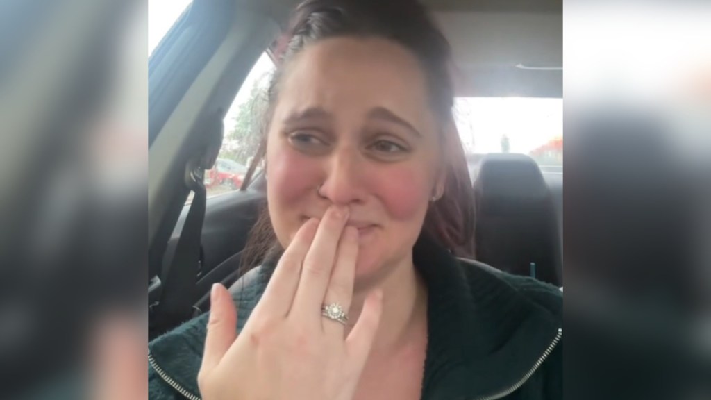 A woman sits in her car as she starts to cry. She holds a hand up to her mouth.