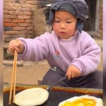 A toddler concentrates as he works to flip bread he's cooking using chopsticks. There's also an egg cooking next to the bread. The toddler is cooking outside.