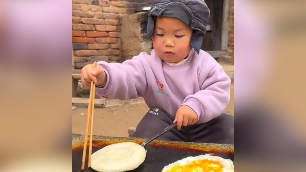 A toddler concentrates as he works to flip bread he's cooking using chopsticks. There's also an egg cooking next to the bread. The toddler is cooking outside.