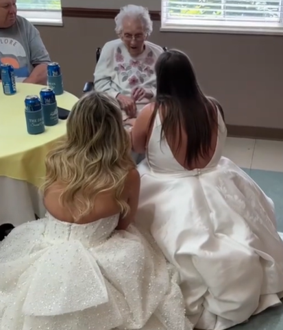 Two brides dressed in white kneeling in front of their great-grandma.