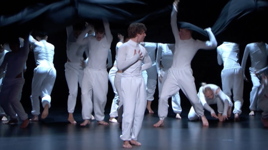 A group of people in all white dance on the "AGT" stage. One person stands in the front while the others scatter from under a large curtain.
