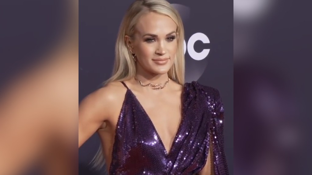 Carrie Underwood poses with a serious expression on a red carpet while in a sparkly dark purple dress.