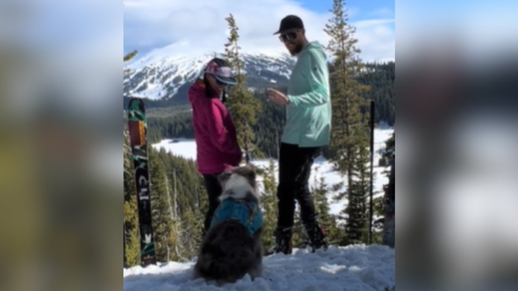 A man and woman stand on a snow-covered cliffside. They smile and laugh as they turn to look back at their dog who is sitting on the ground.