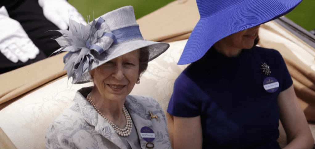 Princess Anne smiles while sitting in the backseat of a car with no roof. She's sitting next to someone whose face is concealed by their hat.