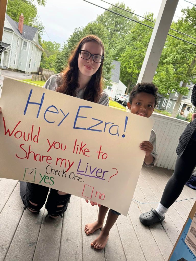 A young woman kneels next to a small child while holding a big sign. 