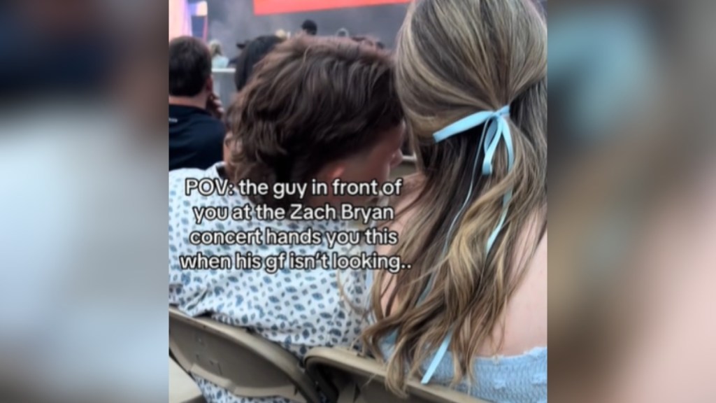 View from behind a man leaning over to the woman next to him, talking. Text on the image reads: POV: the guy in front of you at the Zach Bryan concert hands you this when his gf isn't looking...