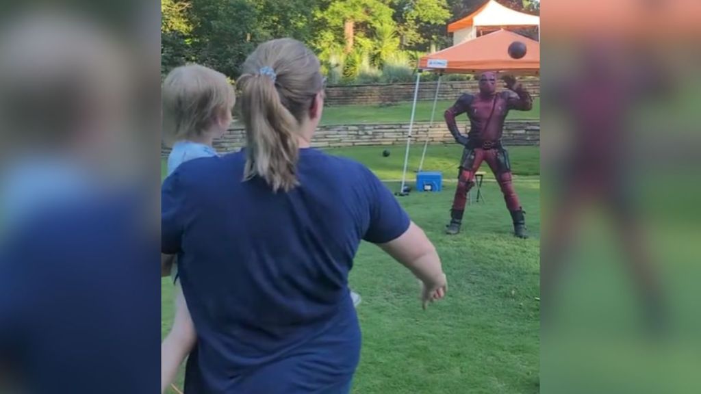 A woman holding a baby and throwing a ball at Deadpool.