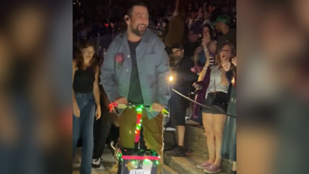 Noah Kahan smiles wide as he rides on a scooter past excited fans