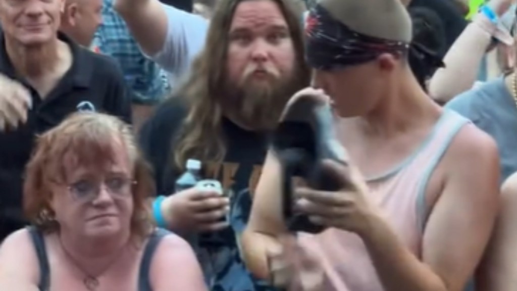 A woman look disappointed as she stands next to fellow concert goers. A tall man next to her looks at her, something in his hand