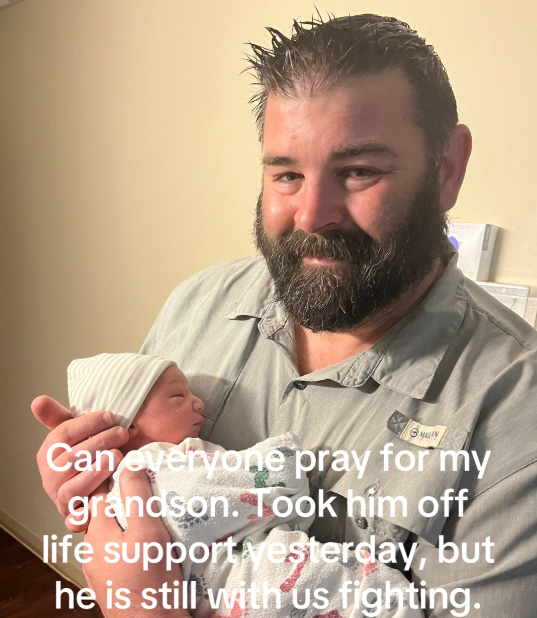 A man gives a small smile while holding a baby. Text on image reads: Can everyone pray for my grandson. Took him off life support yesterday, but he is still with us fighting