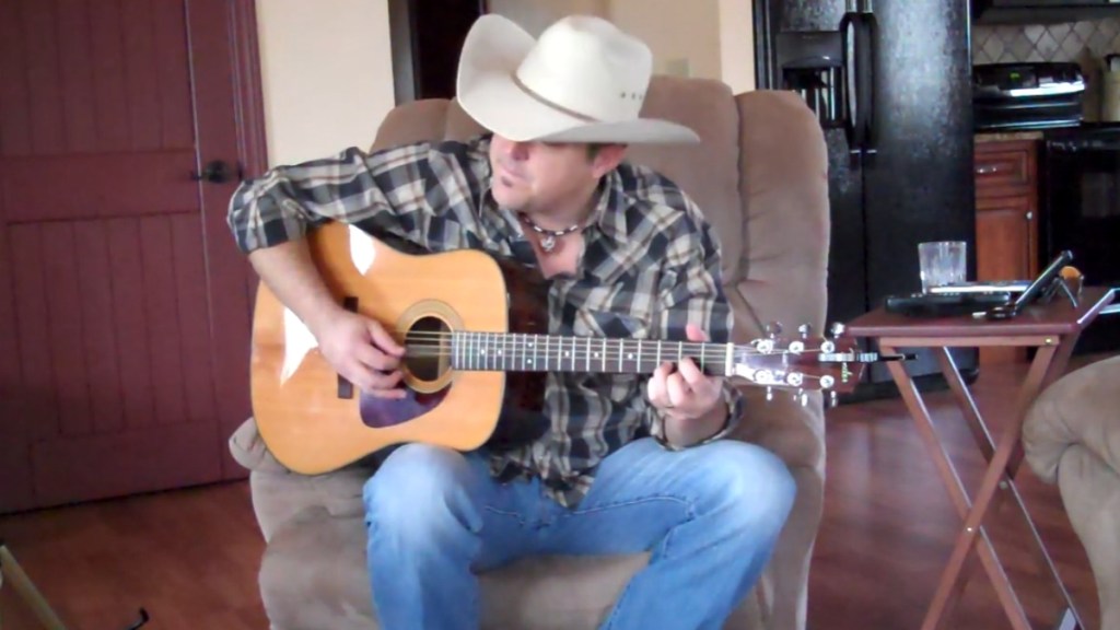 Matt Snook looks away as he sits in a chair and plays the guitar. His large hat mainly covers his face