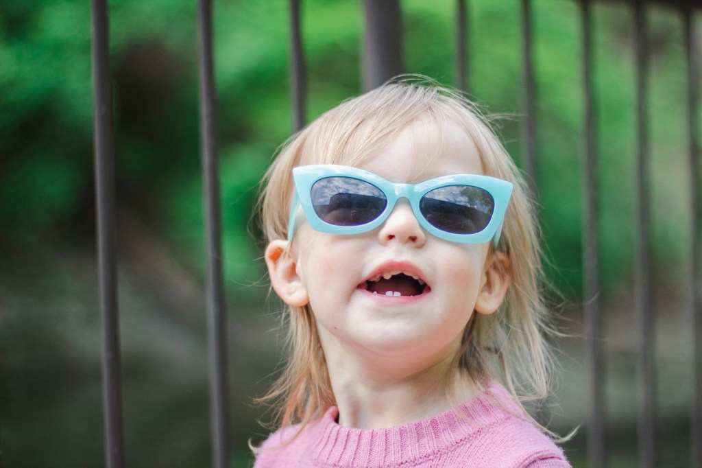 A toddler wearing blue sunglasses and a pink sweater.