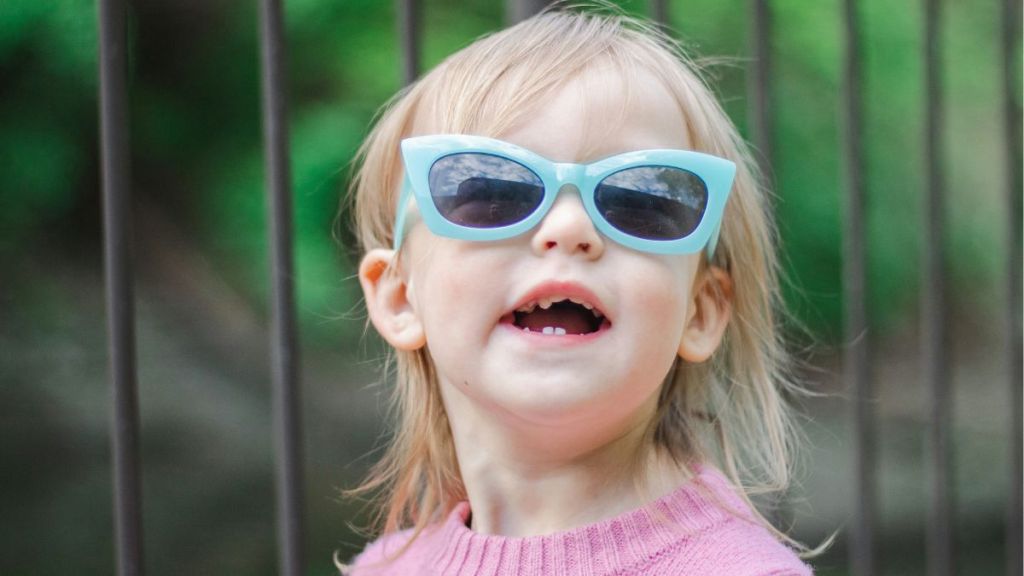 A toddler wearing blue sunglasses and a pink sweater.