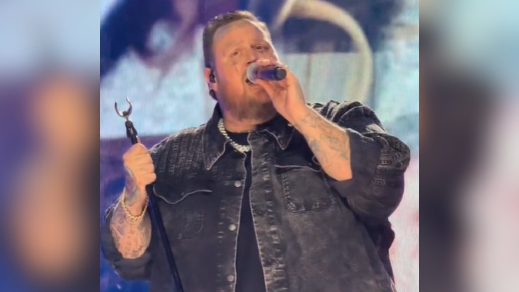 Close up of Jelly Roll singing on stage. He holds a mic stand in one hand and a mic in the other. He looks like he's getting emotional