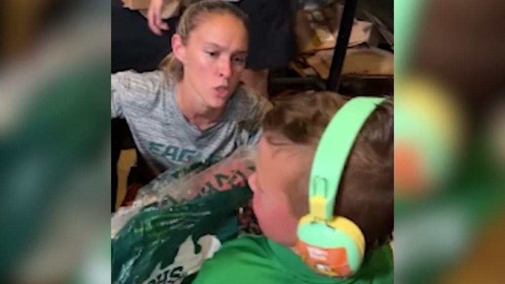 Kylie Kelce squats next to a young boy and talks. The kid is wearing headphones and is holding an Eagles jersey.