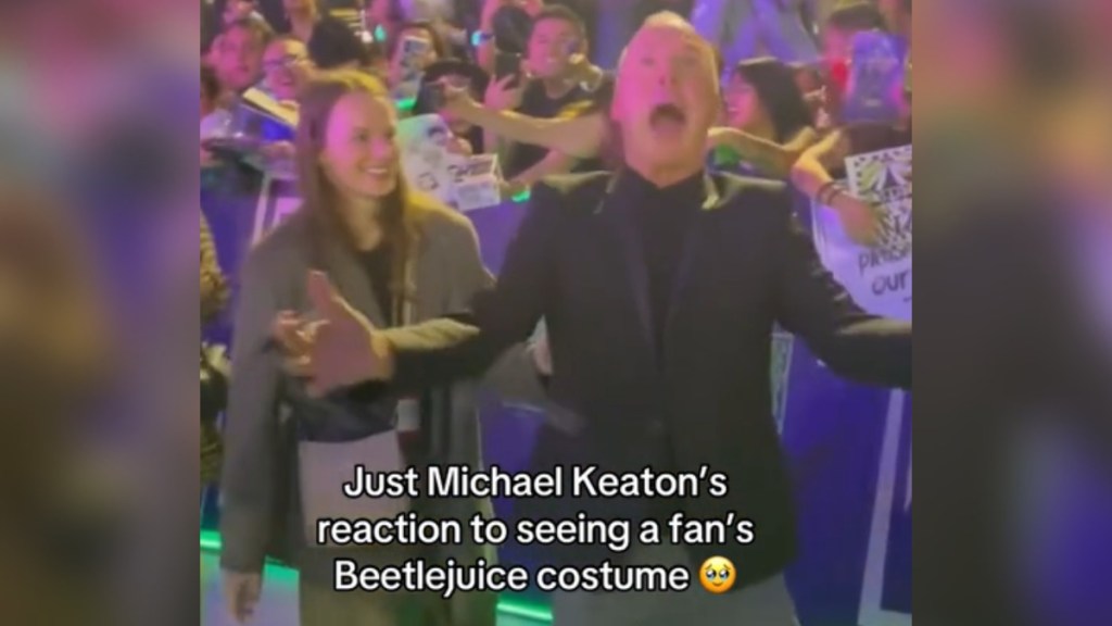 Michael Keaton holds his arms out as he looks amazed, mouth and eyes wide open. A woman stands next to him, smiling as she watches him. Text on the image reads: Just Michael Keaton's reaction to seeing a fan's Beetlejuice costume