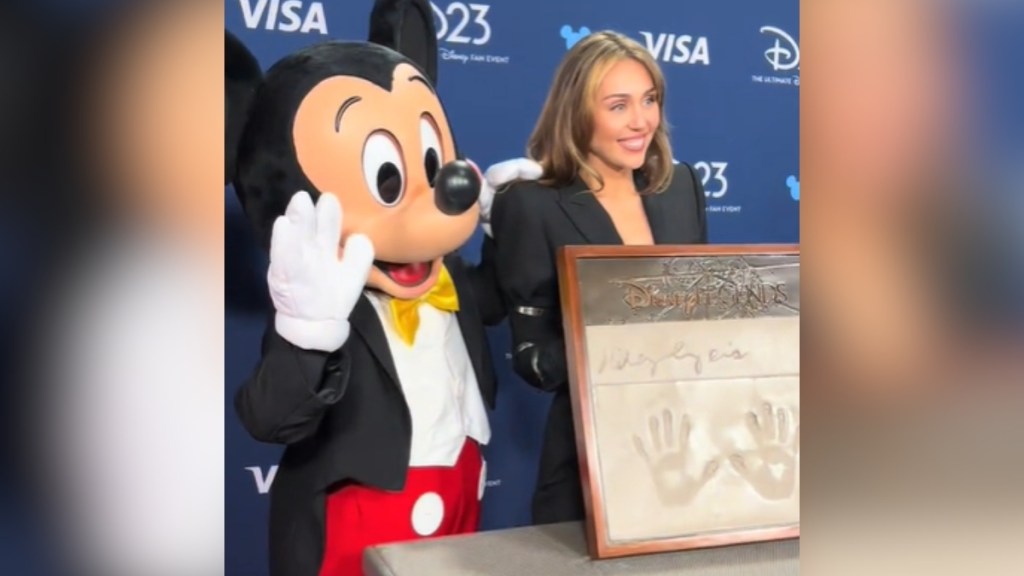 Mickey Mouse waves as he poses with Miley Cyrus. He has an arm around her. Miley is smiling as she holds up cement with hand prints and her signature