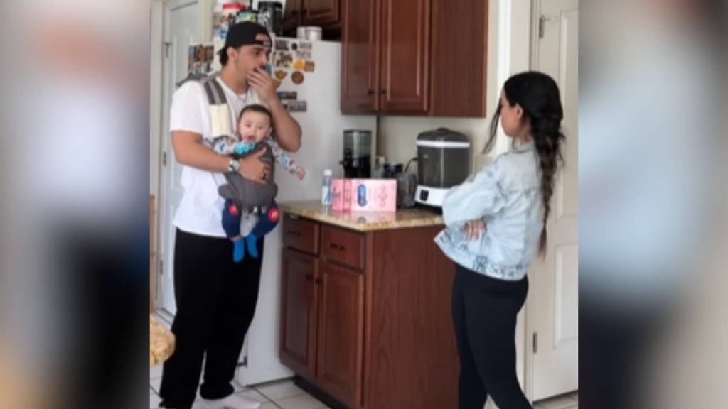 A woman folds her arms, back toward the camera. A man, who has a baby strapped onto his chest, looks at her with eyes wide, one hand covering his mouth. They're in a kitchen