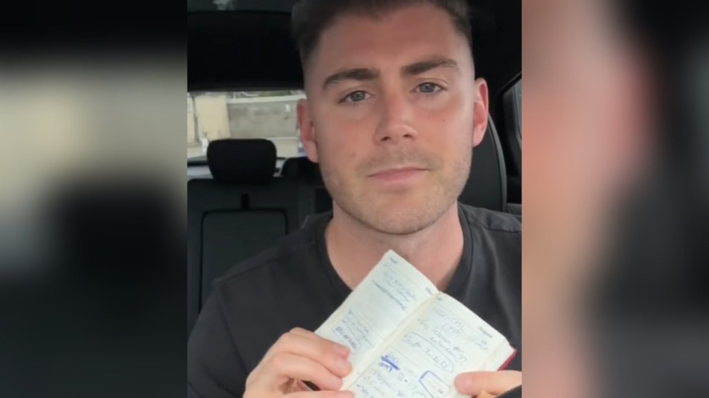 A man looks somber as he holds up a small notebook for the camera. The notebook pages are facing the camera. Written words can be seen on the pages but can't be read from here