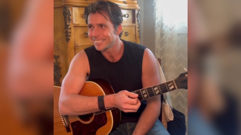 Lukas Nelson looks into the distance, smiling, as he sits with a guitar in his lap