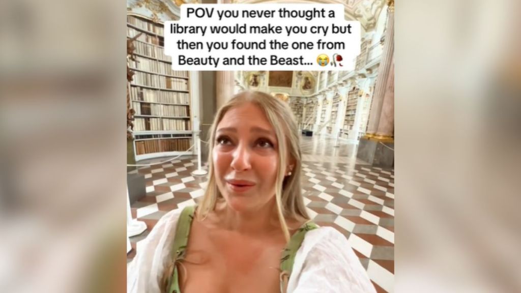 A blonde woman walking through a massive library.