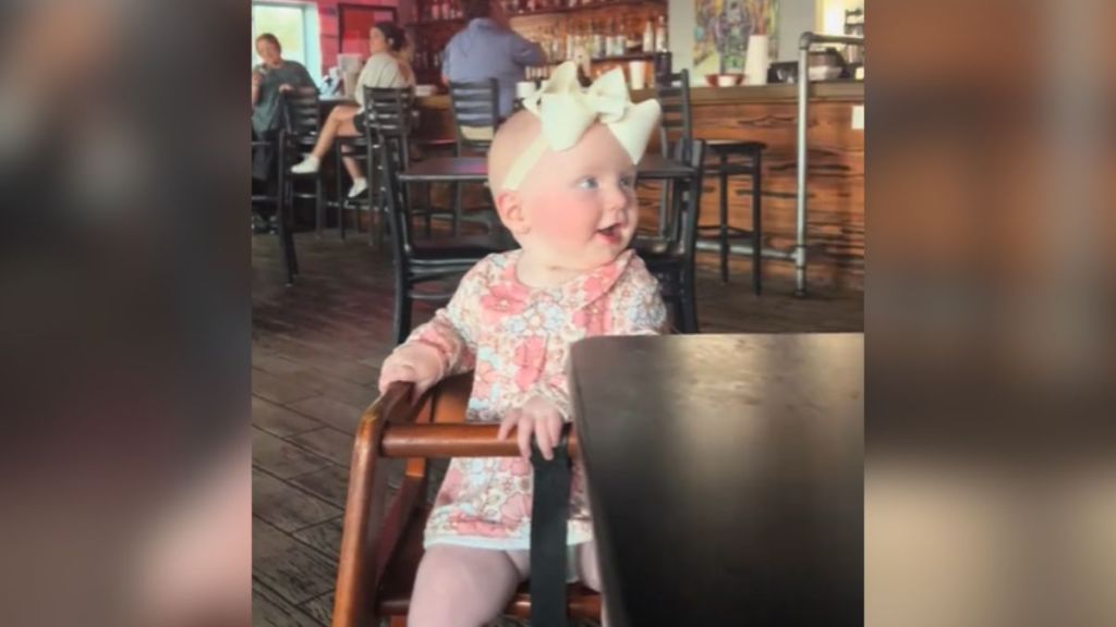 A baby girl with a white bow on her head sitting in a high chair.