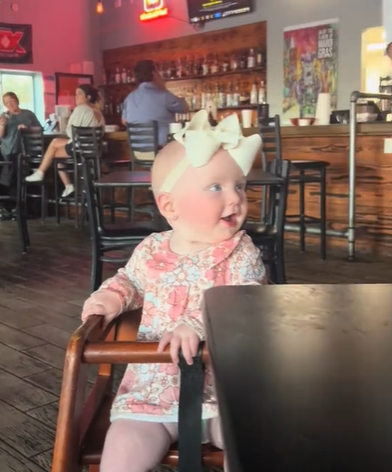 A baby girl with a white bow on her head sitting in a high chair.