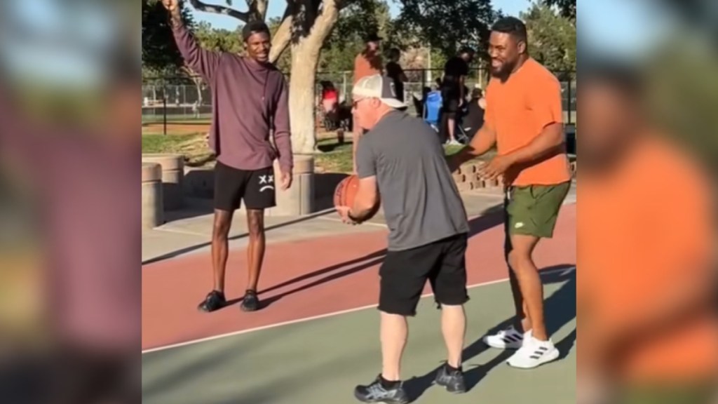 Three man stand on an outdoor basketball court. One holds a basketball while another leans toward him, helping with his form. The third stands nearby, holding something in the air that we can't really see