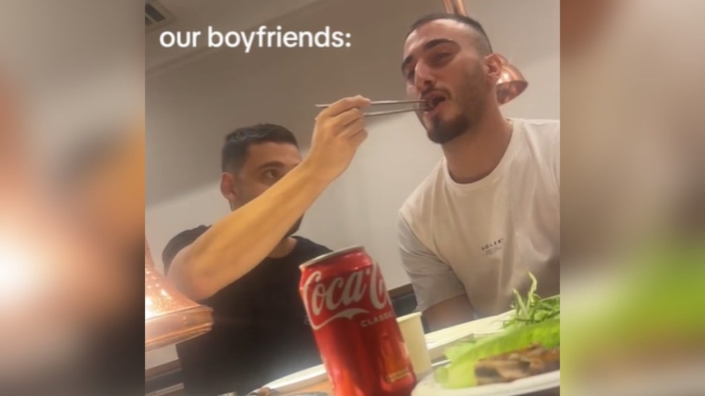 A man reaches over to feed the man sitting next to him, using chop sticks. The second man's mouth is open, ready for the food. Text on the image reads, "Our boyfriends:"