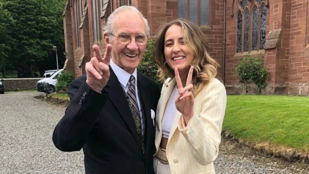 An elderly man, Tommy, and a young woman, Aoife, smile wide as they pose outside. They're both giving peace signs