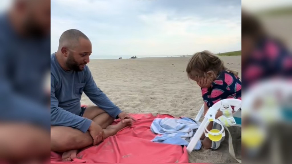 A man sits on a beach towel across from a little girl sitting in a chair. The girl holds her face in her hands, looking sad