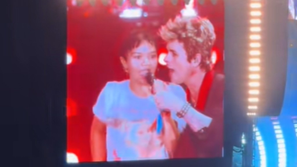 Close up of a large screen at a Green Day concert. On it we see Billie Joe singing into a mic with a young fan