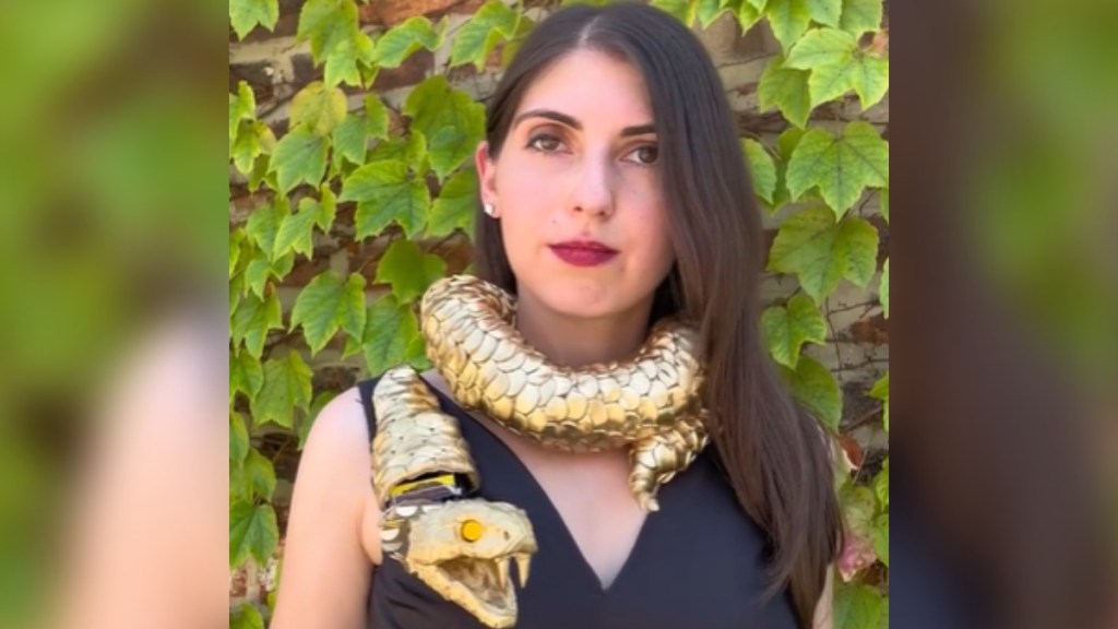 Close up of a woman posing in front of greenery. The top of her dress is black and there's a gold-colored snake wrapped around her neck