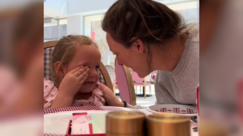 A woman sits with her 5-year-old at a restaurant table. Mom leans close to her daughter and smiles as the little girl rubs away happy tears with one hand