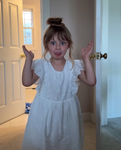A little girl in a white dress making a silly face.