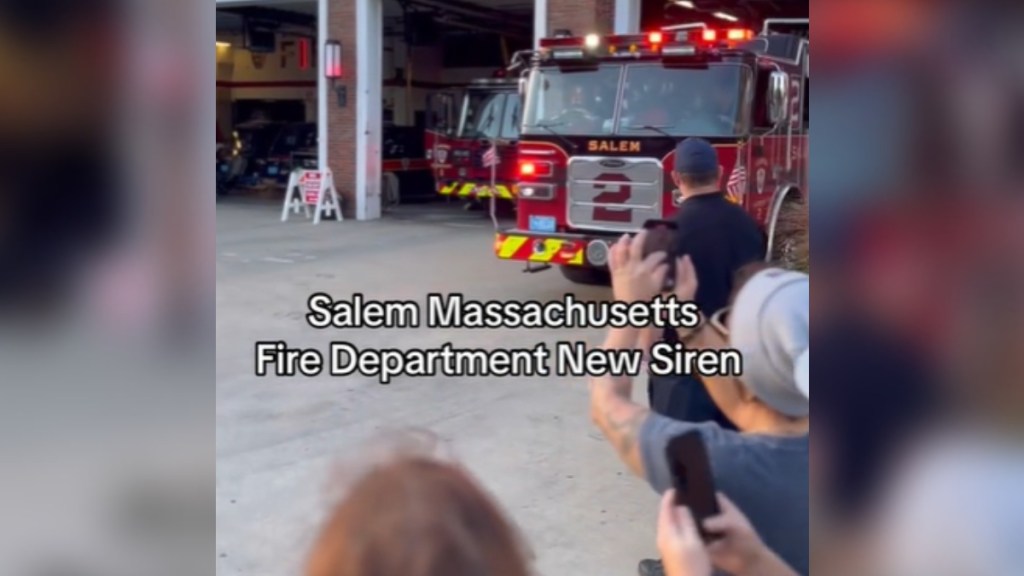 People gather as a firetruck pulls out of the station. Text on the image reads: Salem Massachusetts Fire Department New Siren