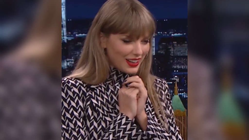 Taylor Swift smiles as she looks down. She's holding her hands up to her chest, waiting for something