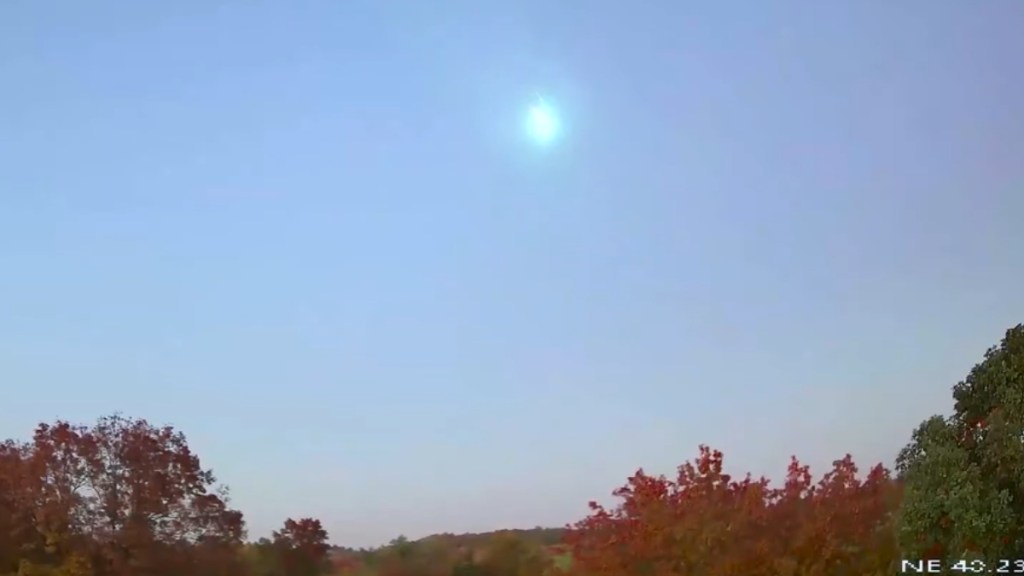 Distant view of a bright meteor flying across the light blue sky. A line of trees can be seen at the bottom of the image