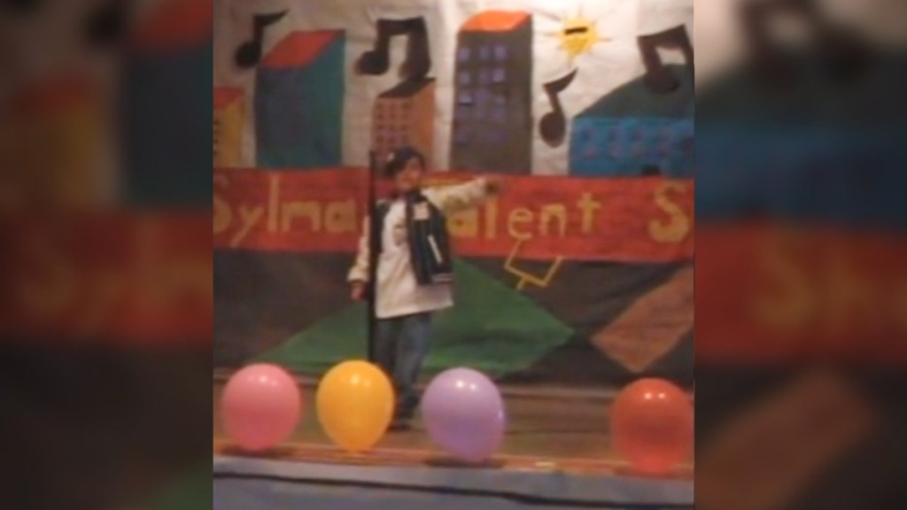 A little kid stands on a stage, singing into a mic. He holds one arm out, pointing out in the audience. Balloons adorn the front of the stage and a colorful banner hangs on the wall behind him