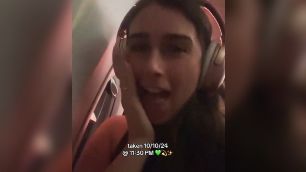 Close up of a woman on a plane. She looks surprised, eyes wide and mouth open, as she holds a hand up to her face. Text on the image reads: taken 10/10/24 @ 11:30 PM