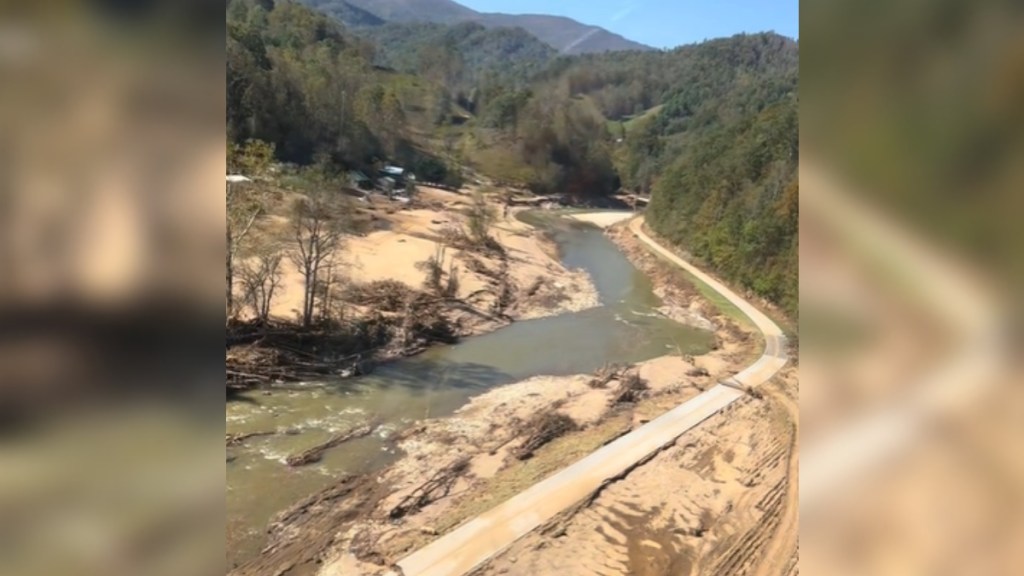 View from a helicopter of devastation left behind from Hurricane Helene in North Carolina. The road is destroyed and the water runs through the muddy area with mountains and trees in the distance