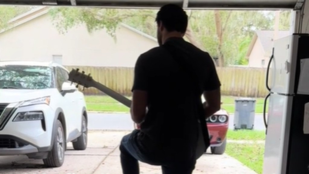 View from behind of a man standing as he plays a guitar. He's inside a garage with the door open