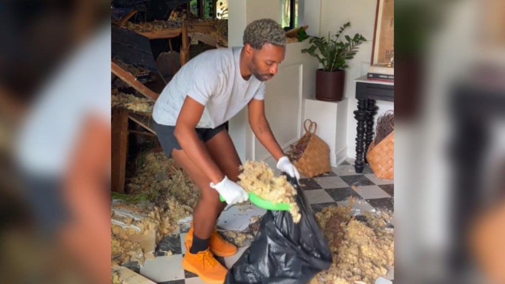A man bends over as he scoops debris into a trash bag. He stands inside his home that is in shambles after Hurricane Helene