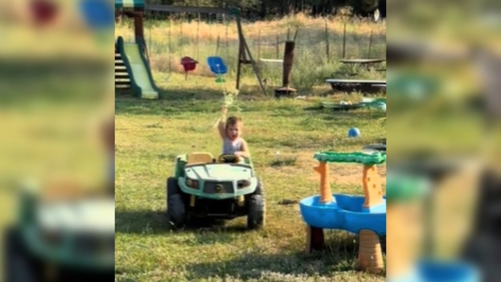 View of a backyard with a playground set and other toys. A little boy rides in a toy tractor, heading toward the camera. He raises an arm in the air, showing off something in his hands
